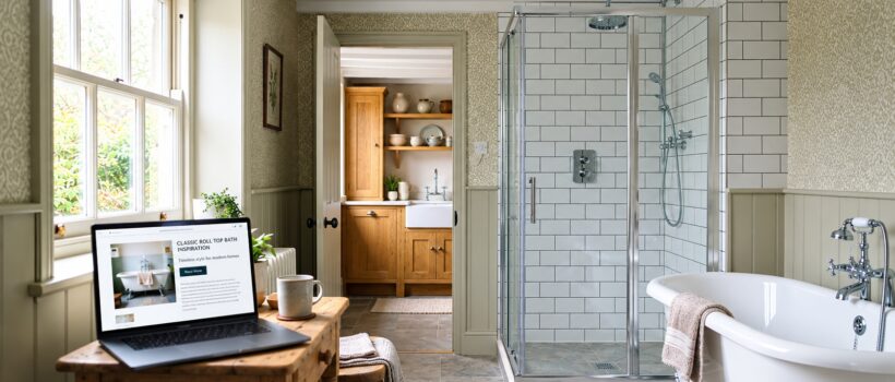 Traditional UK bathroom with subway tiles, roll-top bath and chrome shower, kitchen visible through the doorway, laptop showing a bathroom ecommerce product page